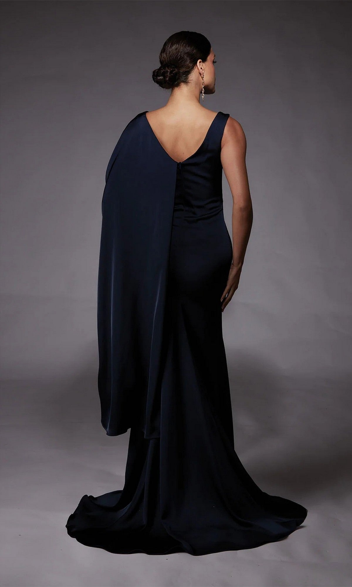 A woman stands facing away against a plain gray background, wearing the Alyce 27722 long formal dress—a floor-length, dark navy one-shoulder gown with a draped cape detail.
